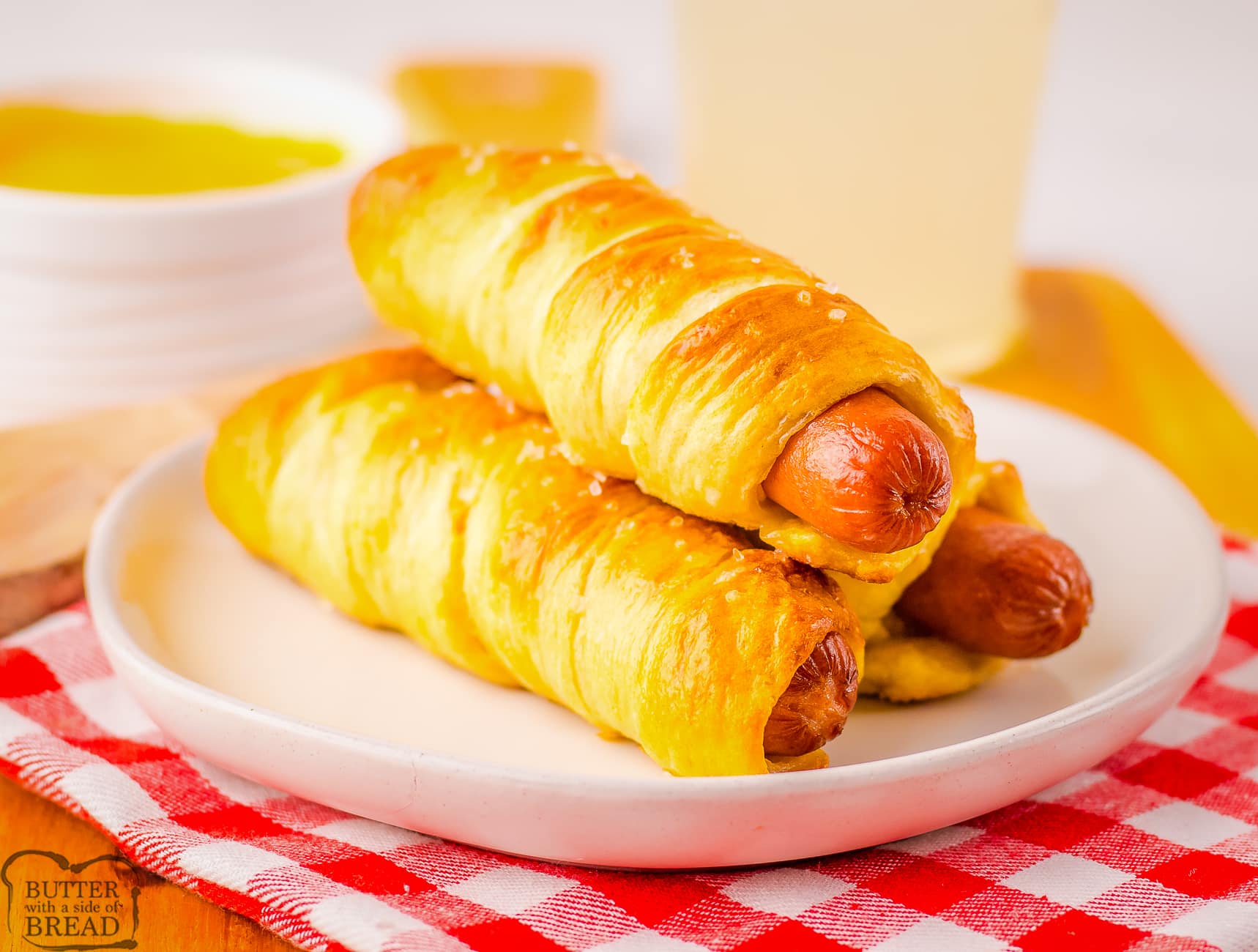 3 pretzel dogs on a white plate with a red checked cloth underneath