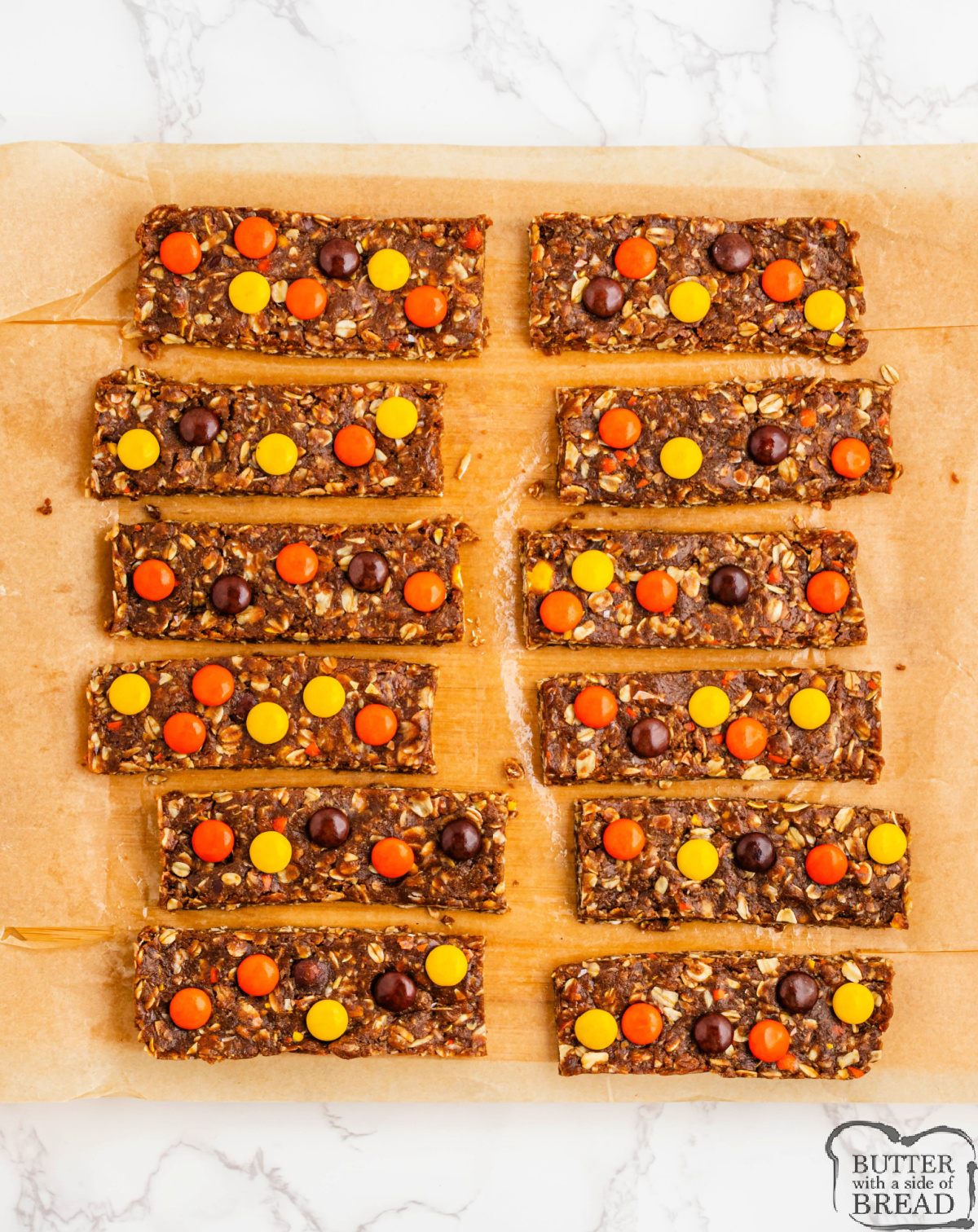 No Bake Reese’s Pieces Protein Bars cut into squares and stacked on parchment paper, showing colorful candy pieces and a chewy peanut butter oat texture.