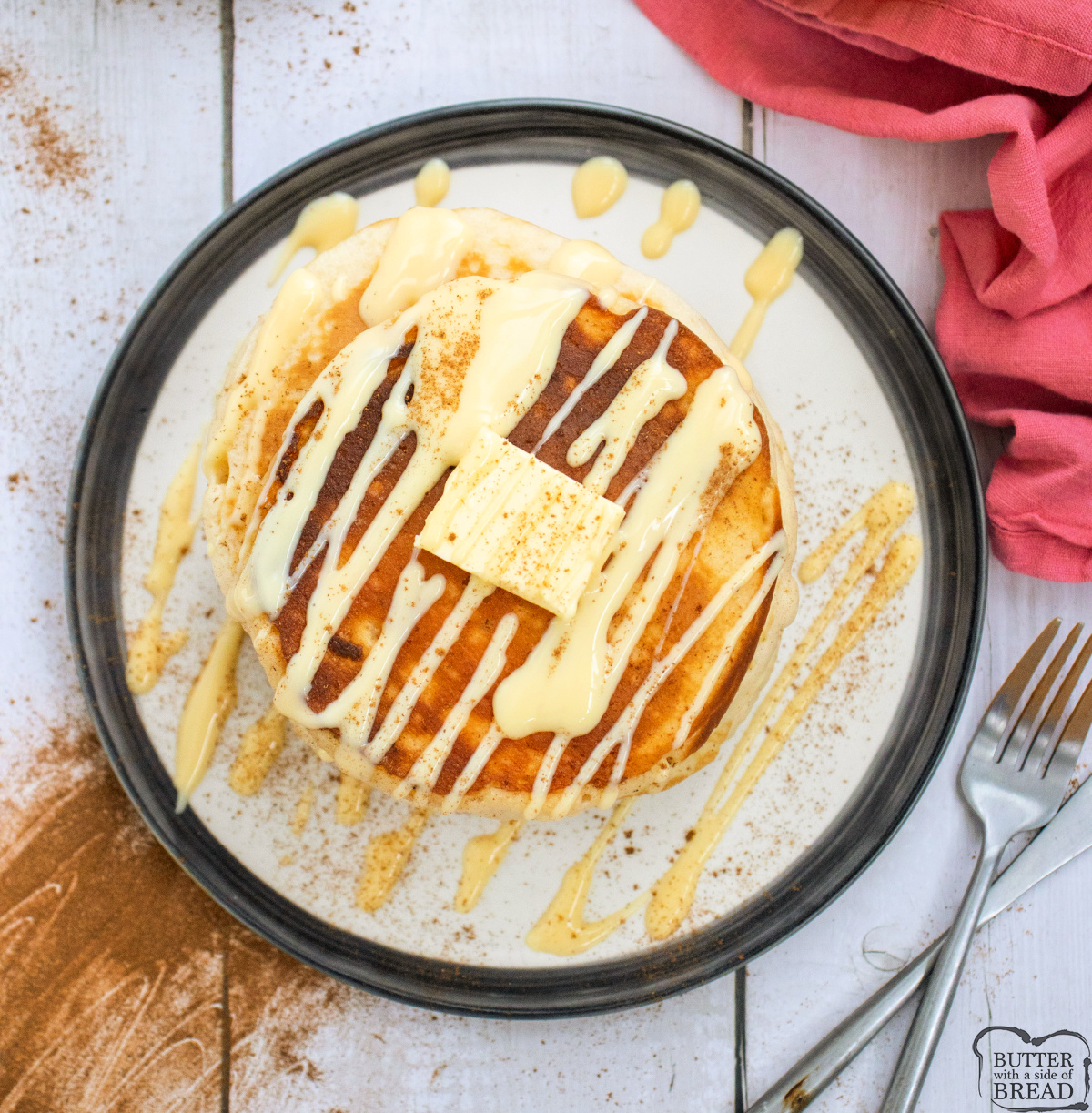 Overhead photo of golden eggnog pancakes topped with butter and drizzled with eggnog glaze, served with cinnamon on a white rustic table.