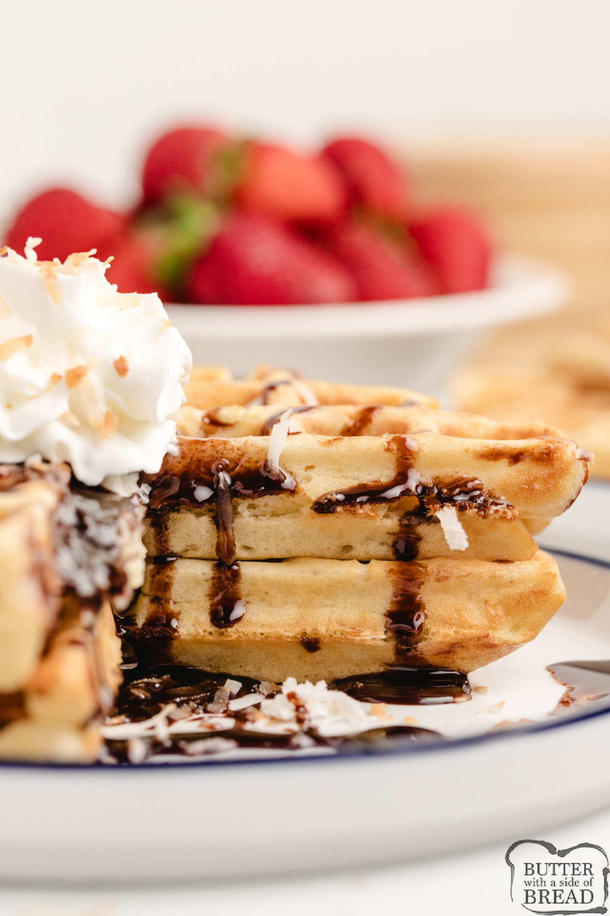 Stack of golden coconut waffles with whipped cream, chocolate syrup, and flaked coconut on top.